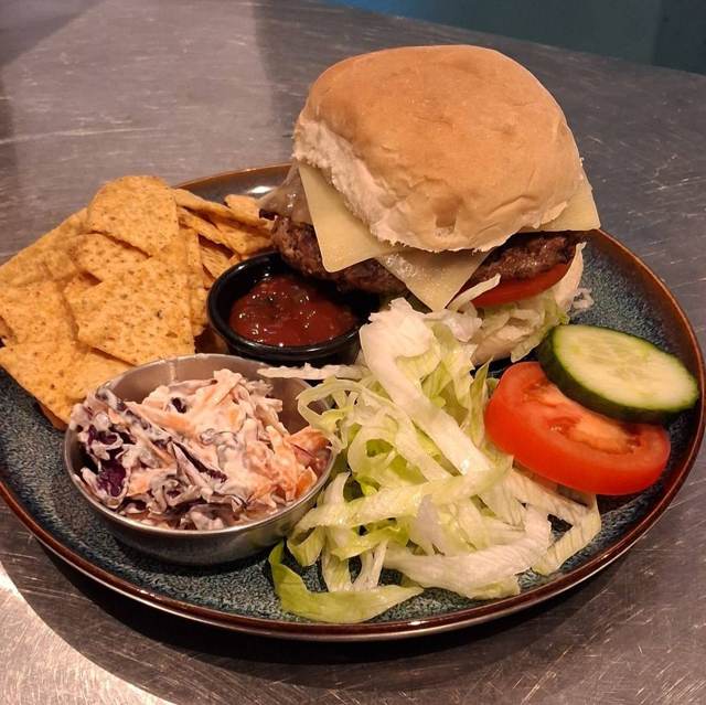 A beef burger with cheese in a flour bap, with nachos, coleslaw, salad and a salsa pot