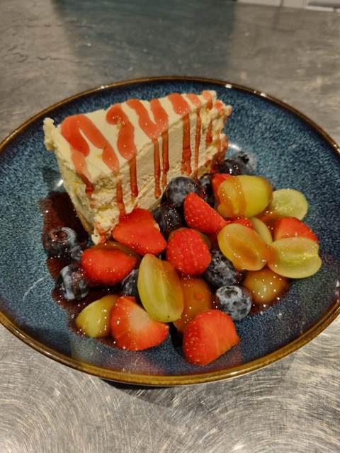 A slice of cheesecake drizzled with berry coulis, served with strawberries, blueberries and grapes