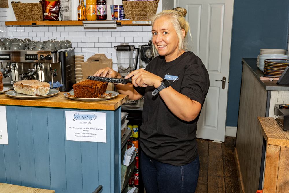 Sammy smiling behind the counter as she slices one of her cakes, with a lemon drizzle loaf beside it